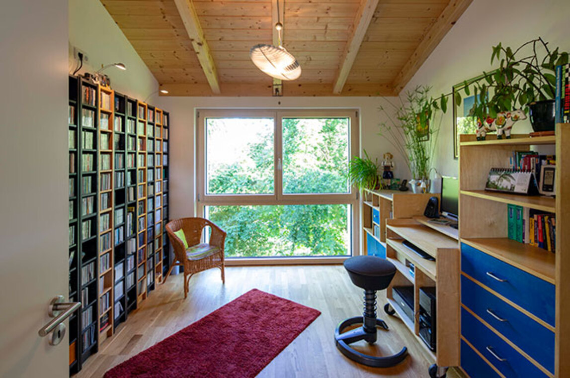 Ein gemütlicher Raum im Passivhaus Bad Aibling zeigt Holzmöbel, ein großes Fenster und üppige Pflanzen.