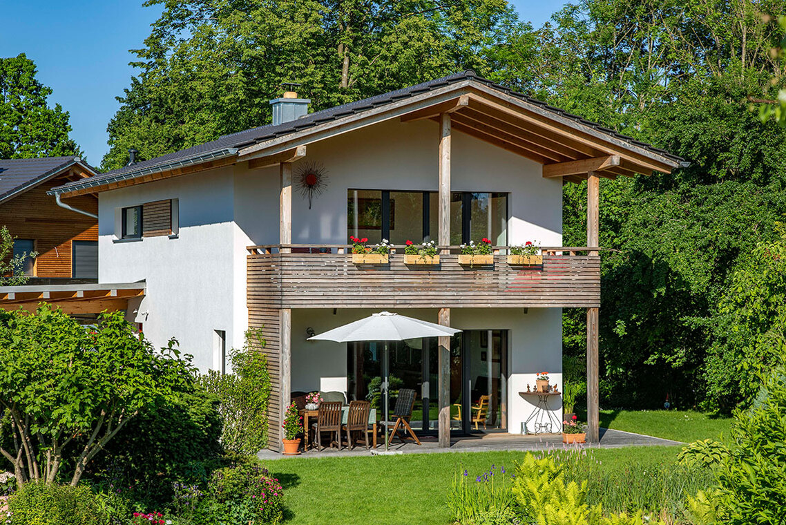 Ein modernes Passivhaus in Bad Aibling mit Balkon und Gartenmöbeln steht umgeben von üppigem Grün im Sonnenschein.