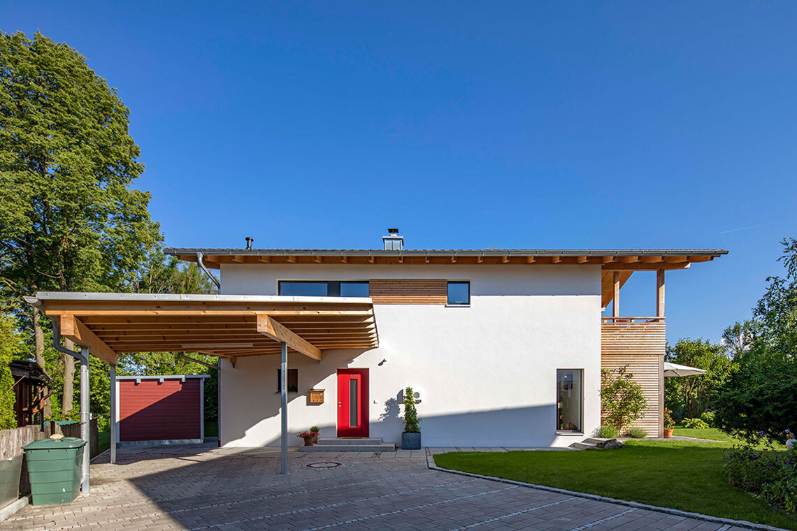 Ein modernes Passivhaus in Bad Aibling mit roter Tür und Carport steht unter klarem, blauem Himmel.