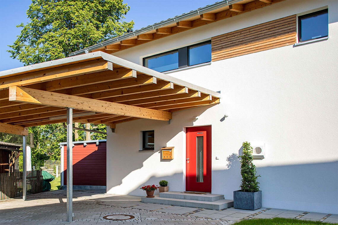 Ein modernes Passivhaus in Bad Aibling mit roter Tür und Holzvordach strahlt im hellen Sonnenlicht.