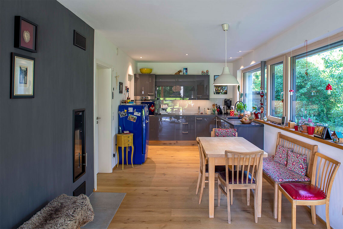 Eine moderne Passivhaus-Küche in Bad Aibling zeigt einen Holzboden, einen blauen Kühlschrank und große Fenster mit Gartenblick.