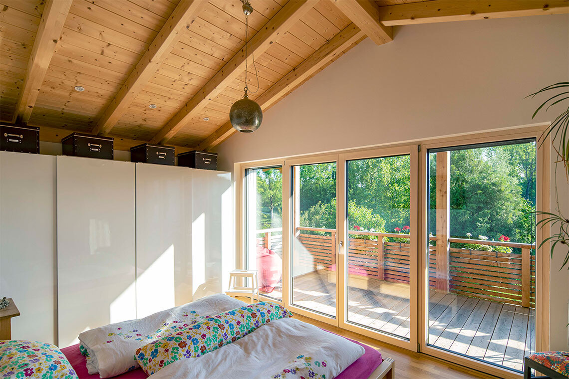 Ein gemütliches Schlafzimmer im Passivhaus mit Holzbalkendecke und großen Fenstern bietet Blick auf einen sonnigen Balkon in Bad Aibling.