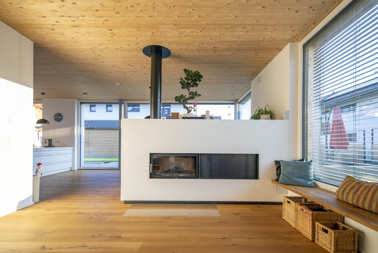 Ein modernes Holzhaus in Oberpframmern präsentiert ein gemütliches Wohnzimmer mit Kamin, Holzboden und großen Fenstern.