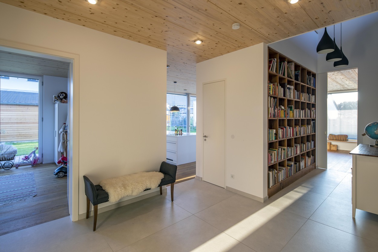 Ein modernes Holzhaus in Oberpframmern zeigt einen hellen Flur mit Bücherregal und angrenzender, lichtdurchfluteter Küche.