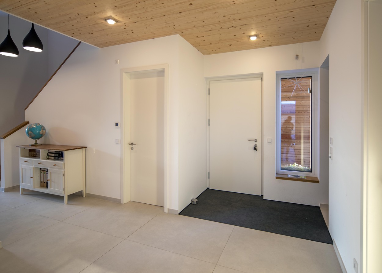 Moderner Eingangsbereich eines Holzhauses in Oberpframmern mit hellen Wänden, Holzdecke und einer kleinen Kommode links.