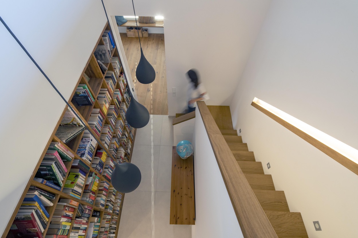 Eine Person steigt in einem modernen Holzhaus in Oberpframmern die Treppe hinunter, vorbei an einer großen Bücherwand.