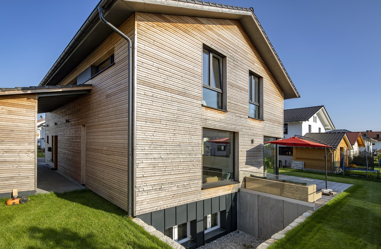 Ein modernes Holzhaus in Oberpframmern mit rotem Sonnenschirm im gepflegten Garten bei strahlendem Sonnenschein.