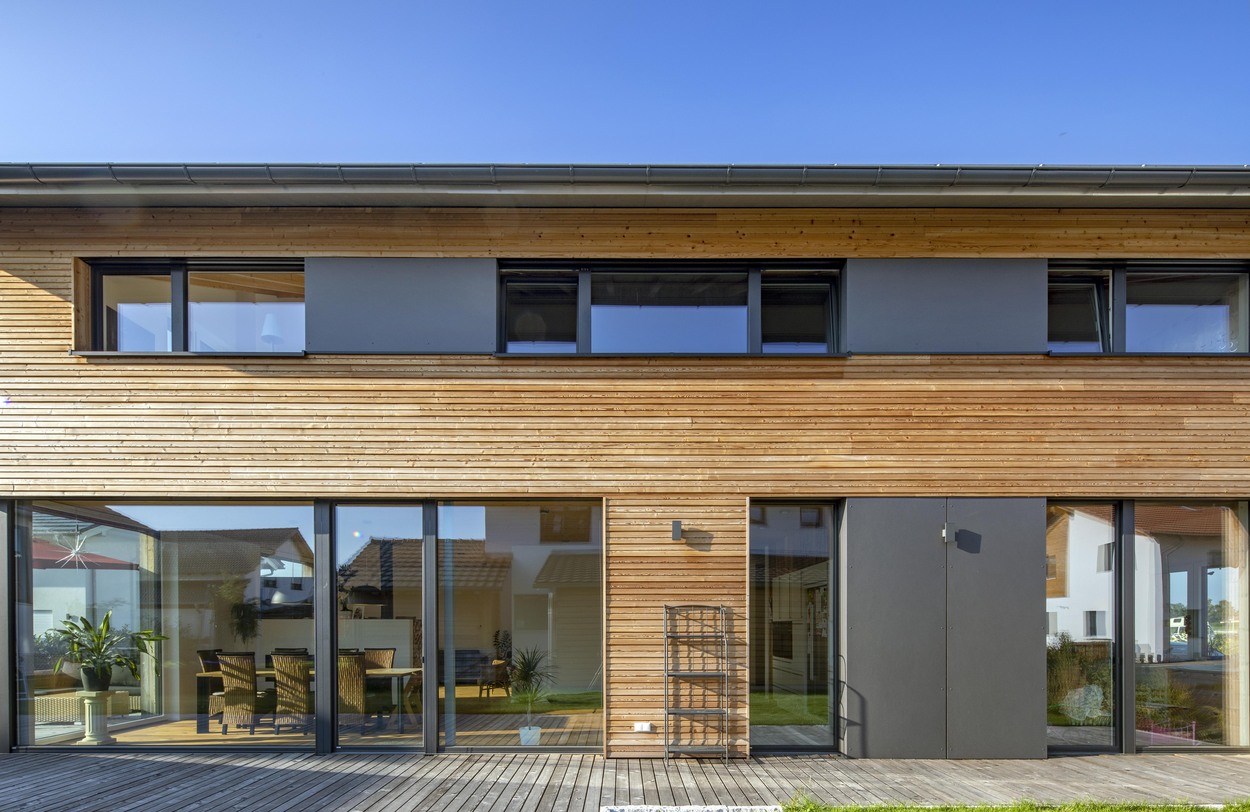 Ein modernes Holzhaus in Oberpframmern mit großen Glasfenstern und einer Terrasse unter strahlend blauem Himmel.