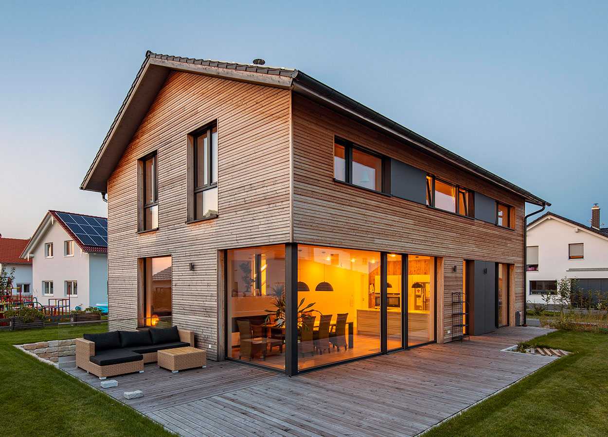 Ein modernes zweistöckiges Holzhaus in Oberpframmern leuchtet bei Abenddämmerung mit großen Fenstern und Terrasse.