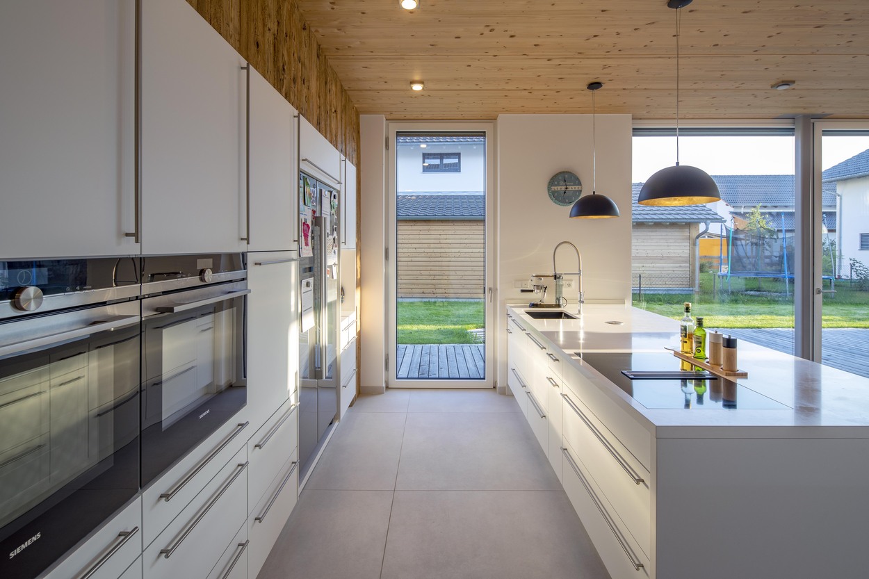 Moderne Küche in einem Holzhaus in Oberpframmern mit Insel, großen Fenstern und viel Tageslicht auf eleganten Oberflächen.