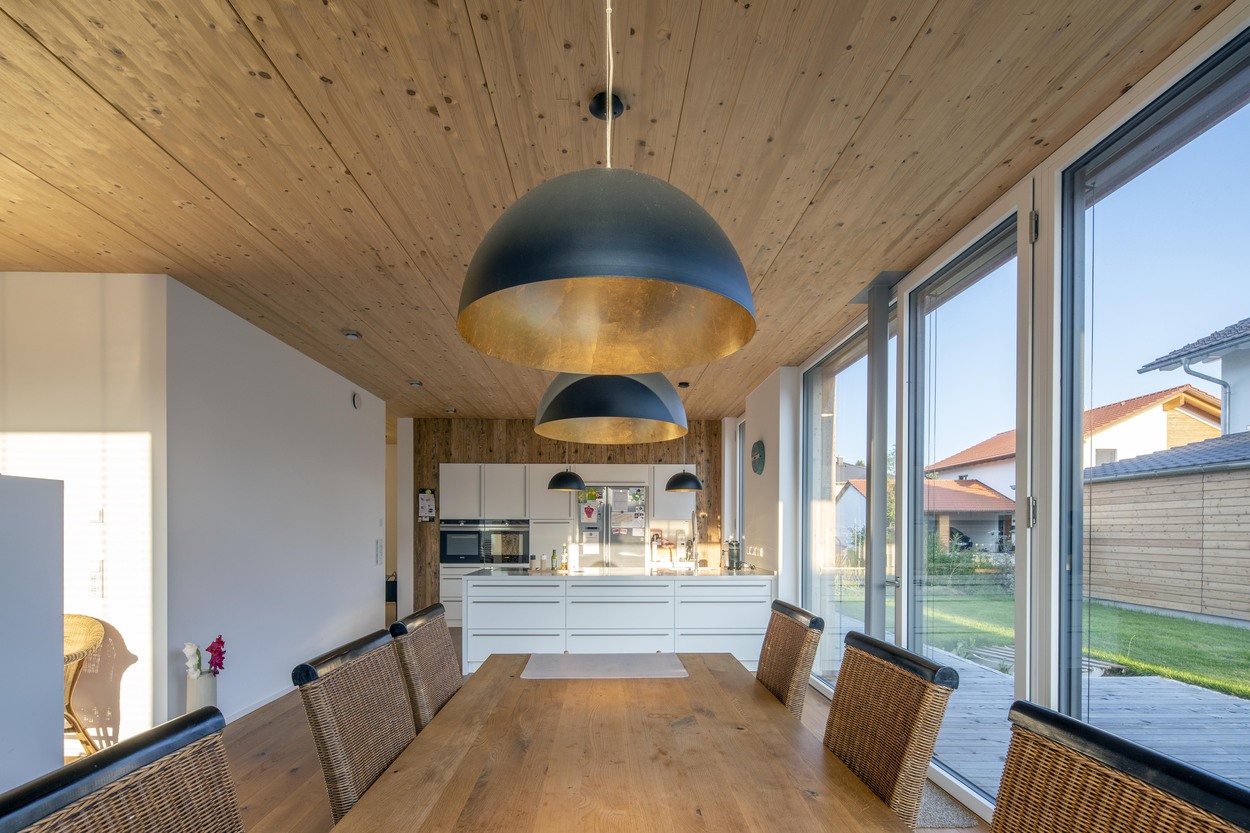 Ein modernes Esszimmer in einem Holzhaus in Oberpframmern mit großen Fenstern und stilvollen Hängelampen über einem Holztisch.