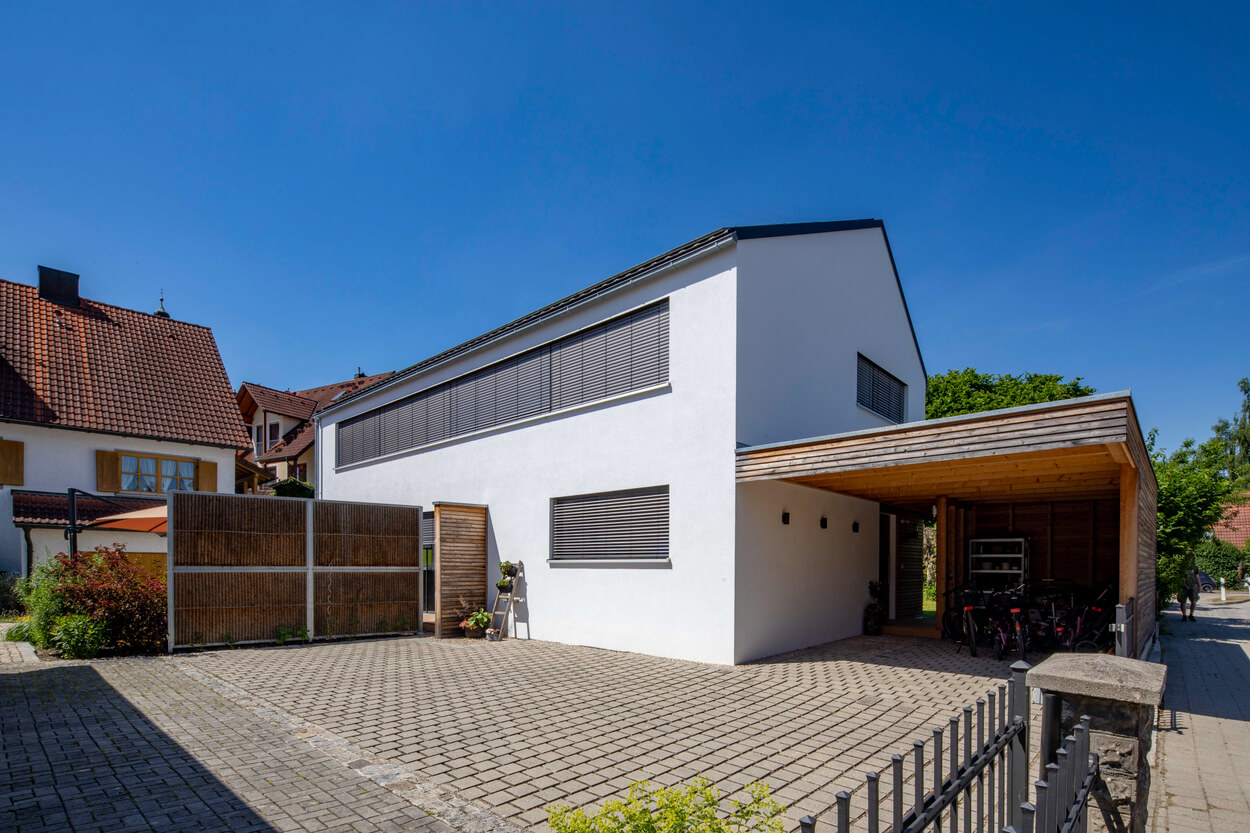 Ein modernes Passivhaus in Fahrenzhausen mit weißer Fassade und überdachtem Carport strahlt an einem sonnigen Tag.