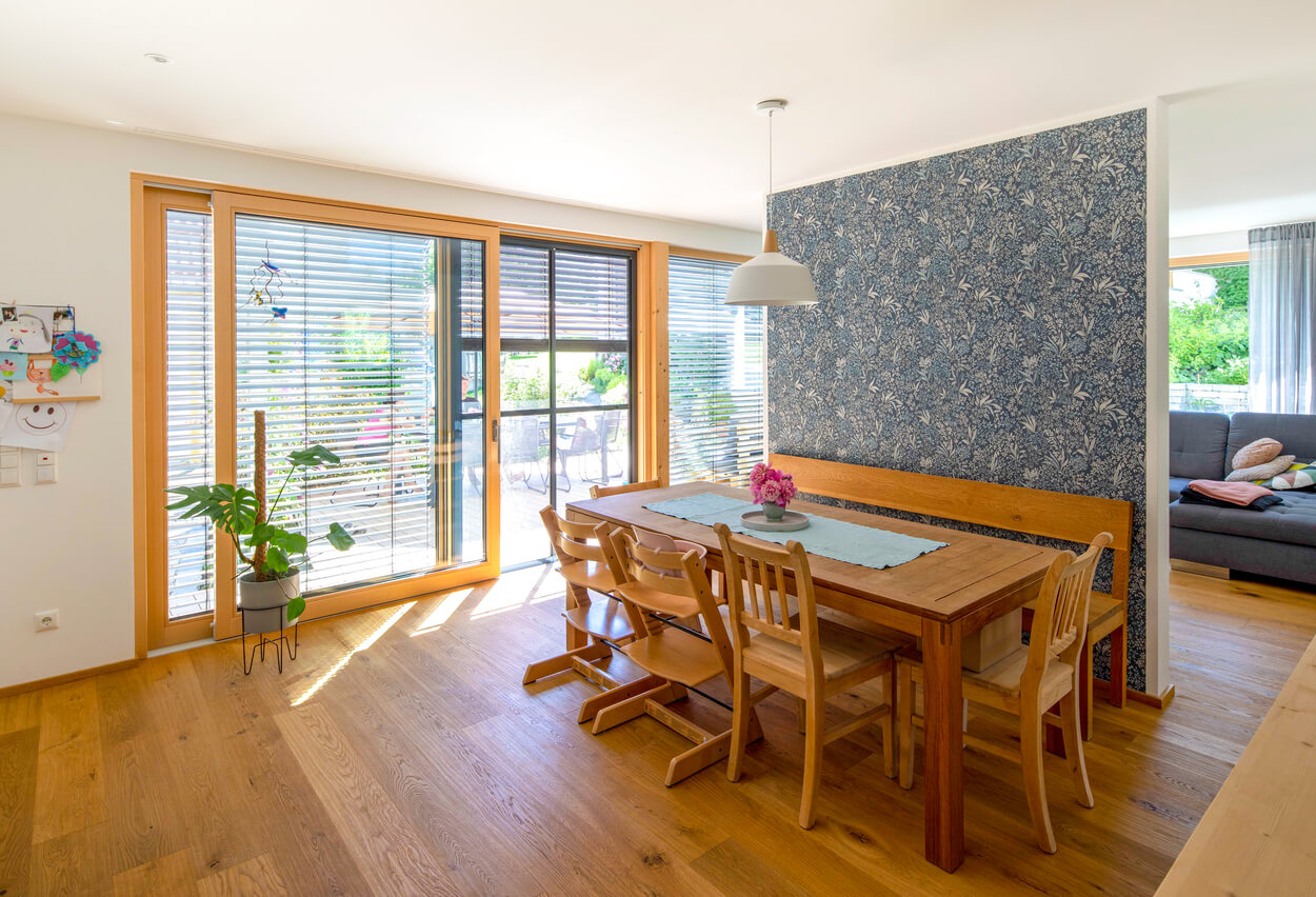 Ein helles Esszimmer im Passivhaus Fahrenzhausen zeigt Holzmöbel, große Fenster und eine stilvolle Tapete.