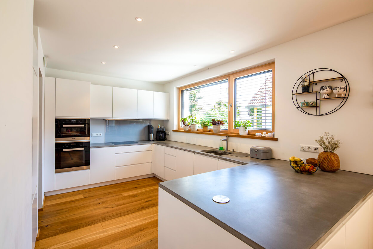 Moderne Küche in einem Passivhaus in Fahrenzhausen mit weißen Schränken, Holzfußboden und großen Fenstern voller Pflanzen.