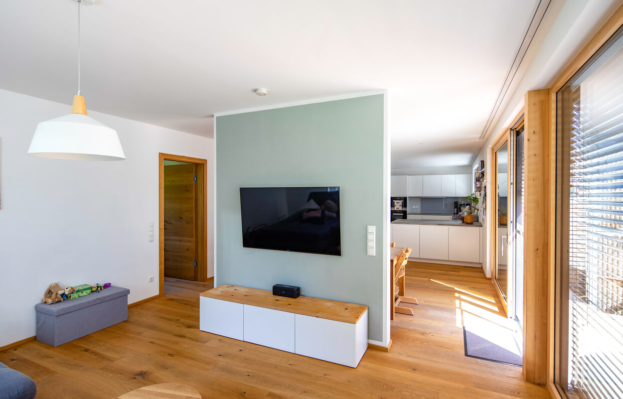 Ein modernes Wohnzimmer im Passivhaus Fahrenzhausen mit Holzfußboden, TV-Wand und Blick in die helle Küche.