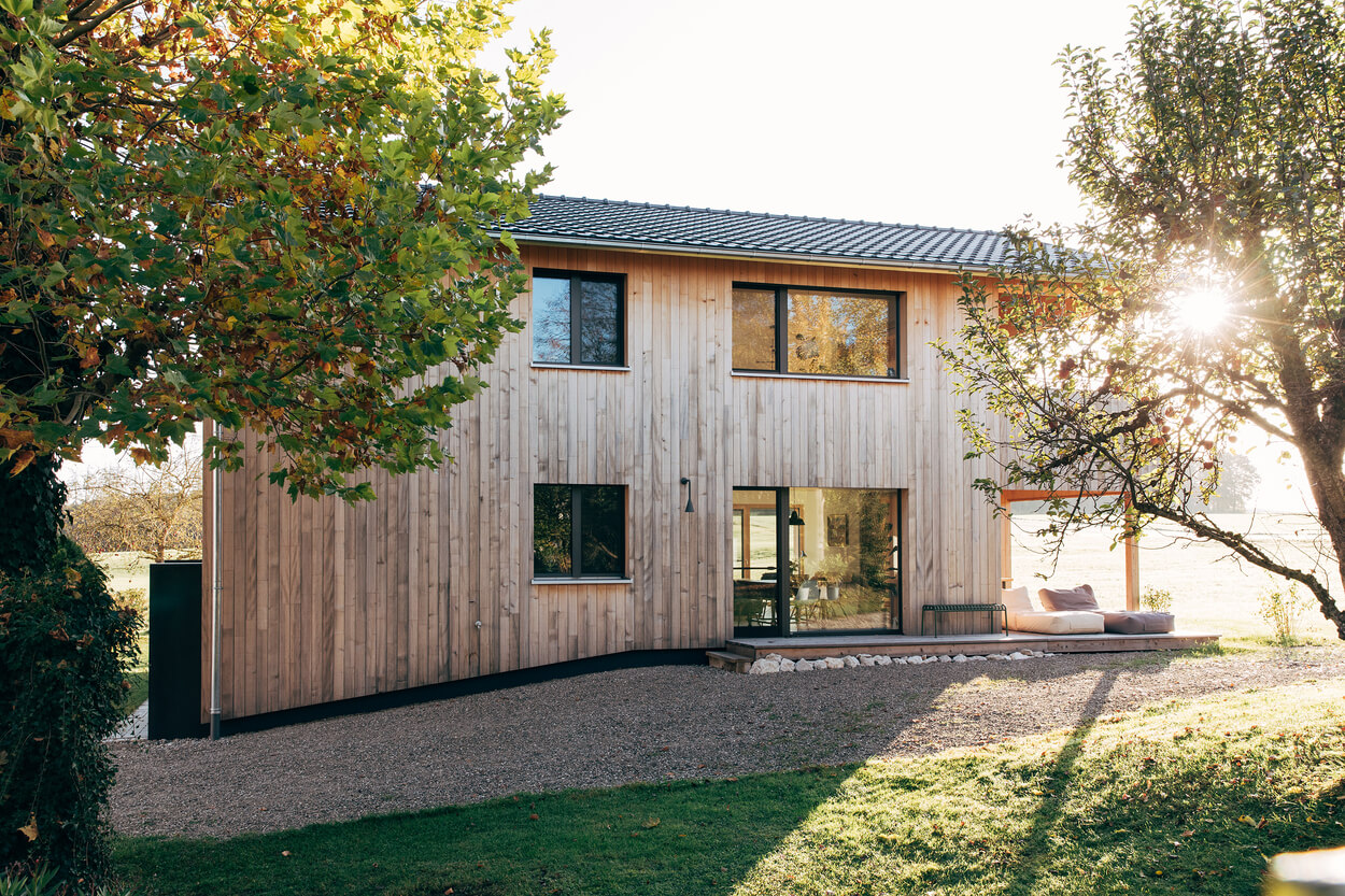 Ein modernes Holzhaus im Chiemgau wird von Sonnenlicht durchflutet, das durch die umgebenden Bäume strahlt.