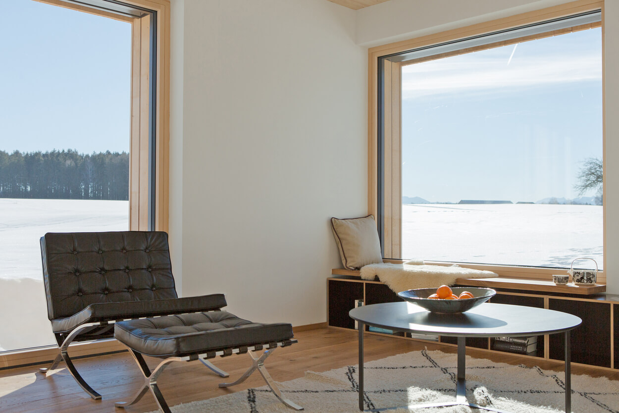 Ein elegantes Wohnzimmer im Holzhaus bietet durch große Fenster einen beeindruckenden Ausblick auf die verschneite Landschaft im Chiemgau.