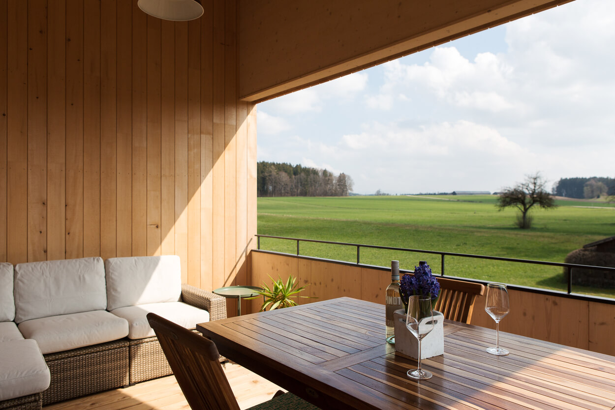 Ein gemütliches Holzhaus im Chiemgau bietet von der Terrasse mit einem Tisch, Wein und Gläsern einen Blick auf grüne Wiesen.