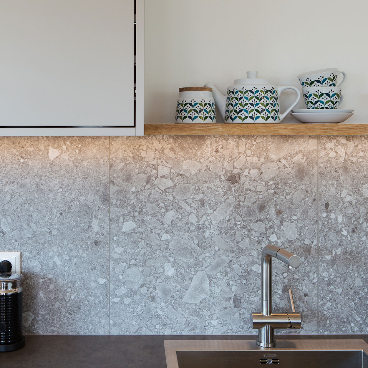 Ein stilvolles Küchenregal im Holzhaus zeigt Geschirr mit blau-grünem Muster vor einer eleganten Steinwand im Ferienhaus Chiemgau.