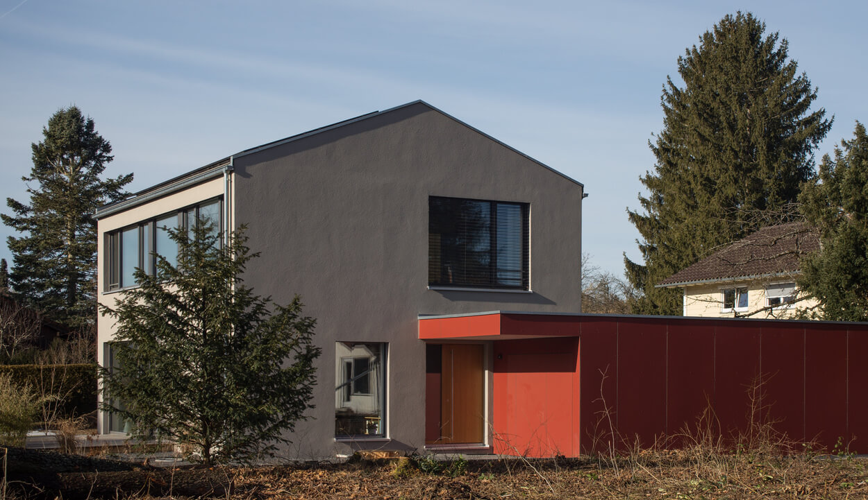 Ein modernes Passivhaus in Baierbrunn mit grauer Fassade und rotem Anbau steht neben hohen Bäumen im Freien.