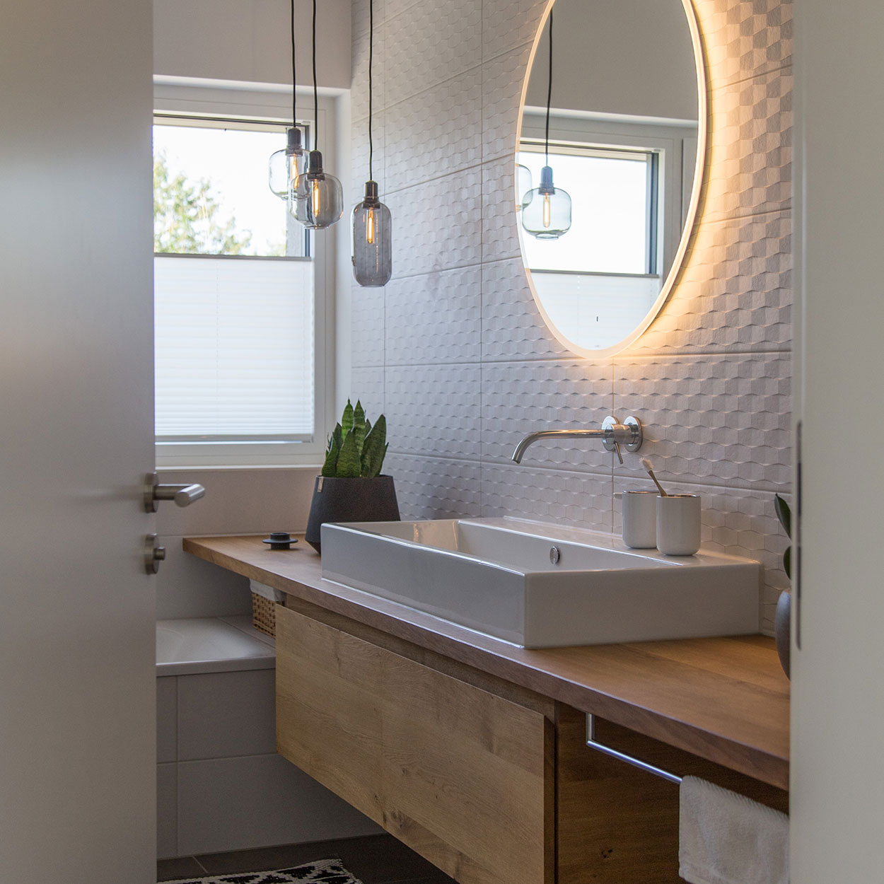 Ein modernes Badezimmer in Baierbrunn zeigt ein beleuchtetes, rundes Spiegel über einem Waschbecken mit Holzablage.