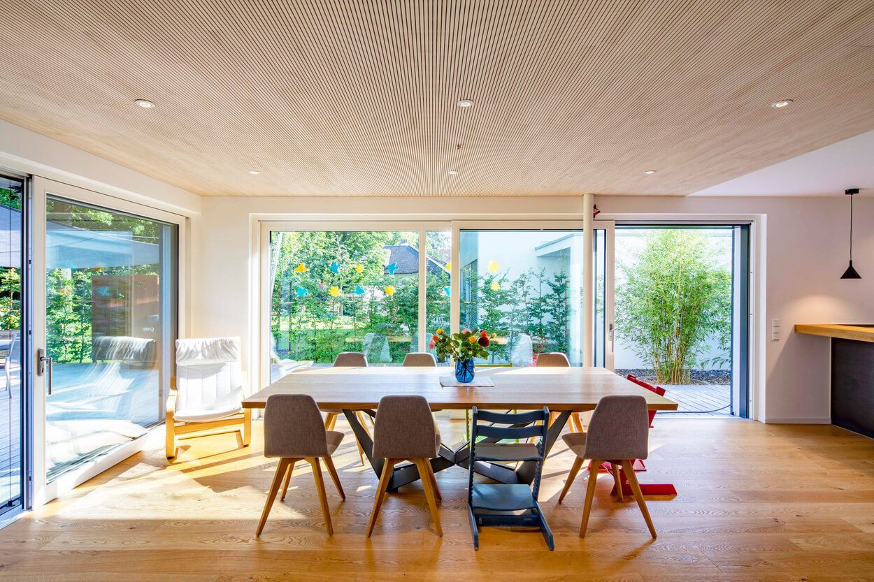 Ein modernes Esszimmer im Passivhaus Eichenau mit großem Holztisch und Blick auf den sonnigen Garten durch große Fenster.