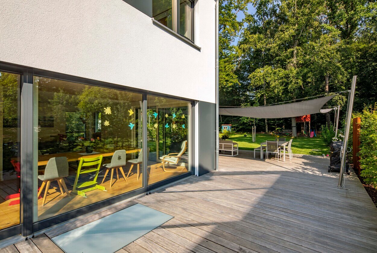 Ein modernes Passivhaus in Eichenau präsentiert eine sonnige Holzterrasse mit gemütlicher Sitzgelegenheit im Garten.