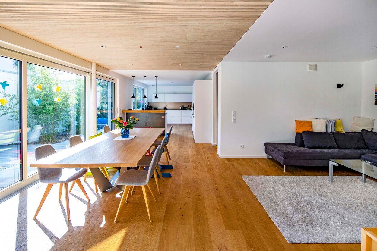 Ein moderner Wohnbereich im Passivhaus Eichenau mit Holzboden, großem Esstisch und gemütlicher Sitzecke lädt zum Verweilen ein.