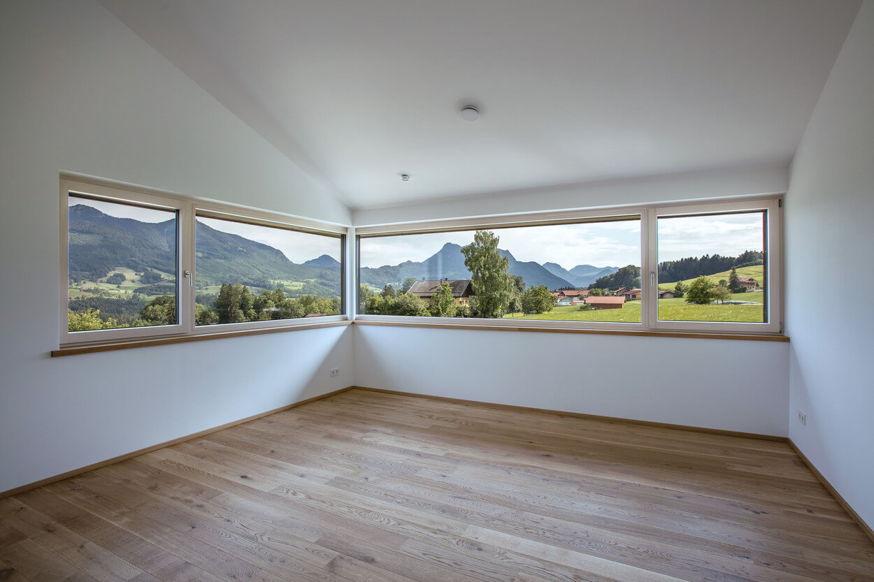 Ein leeres Zimmer mit Holzboden enthüllt durch große Fenster einen atemberaubenden Blick auf die Samerberger Berge.