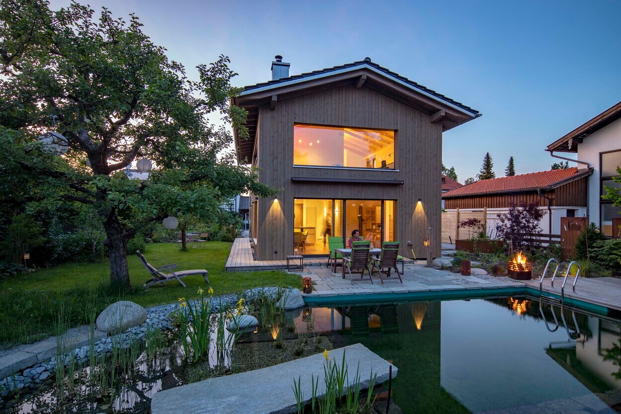 Ein modernes Holzhaus in Otterfing leuchtet bei Abenddämmerung, umgeben von einem beleuchteten Garten und Naturteich.