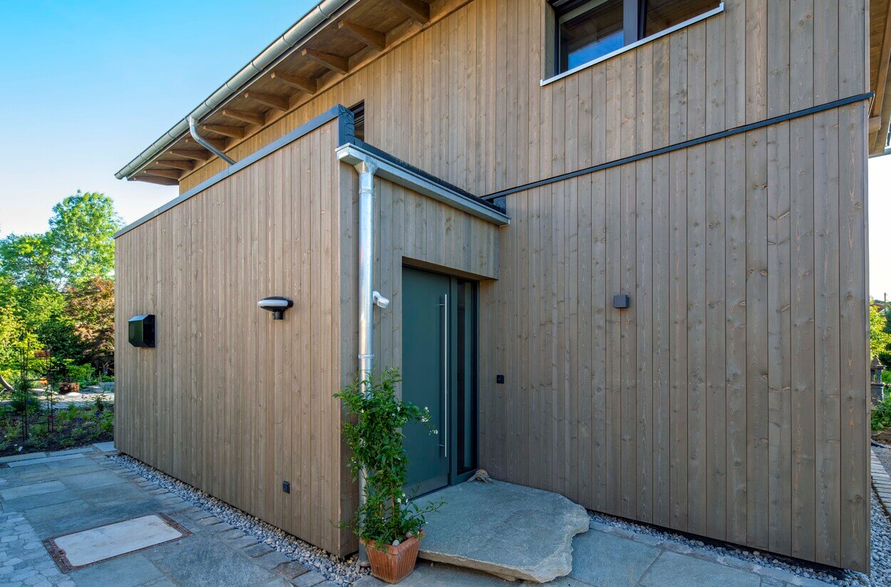 Modernes Holzhaus in Otterfing mit klarer Architektur und begrüntem Eingangsbereich bei sonnigem Wetter.