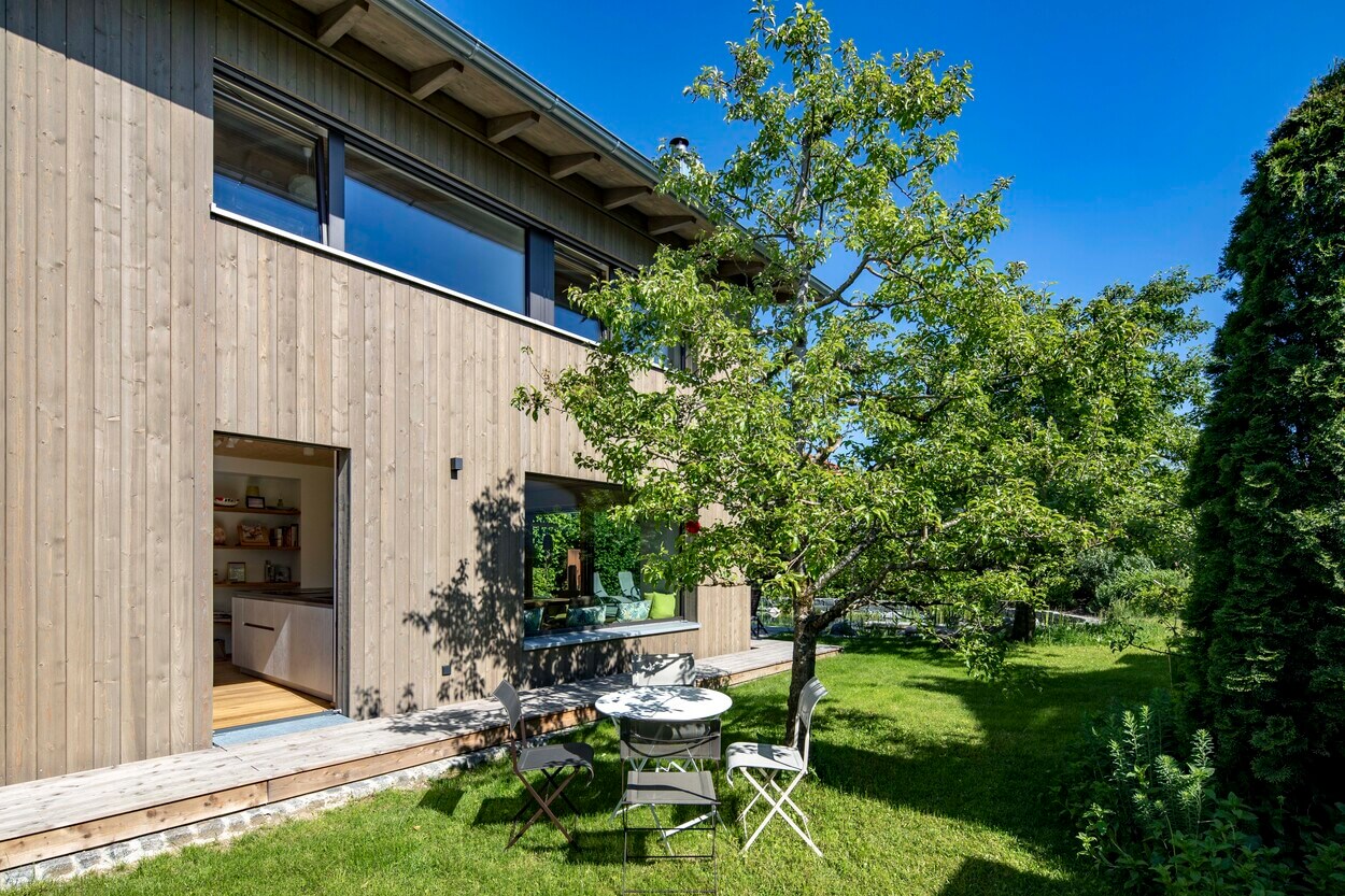 Moderne Außenansicht eines Holzhauses in Otterfing, umgeben von einem grünen Garten mit kleinem Tisch unter einem Baum.