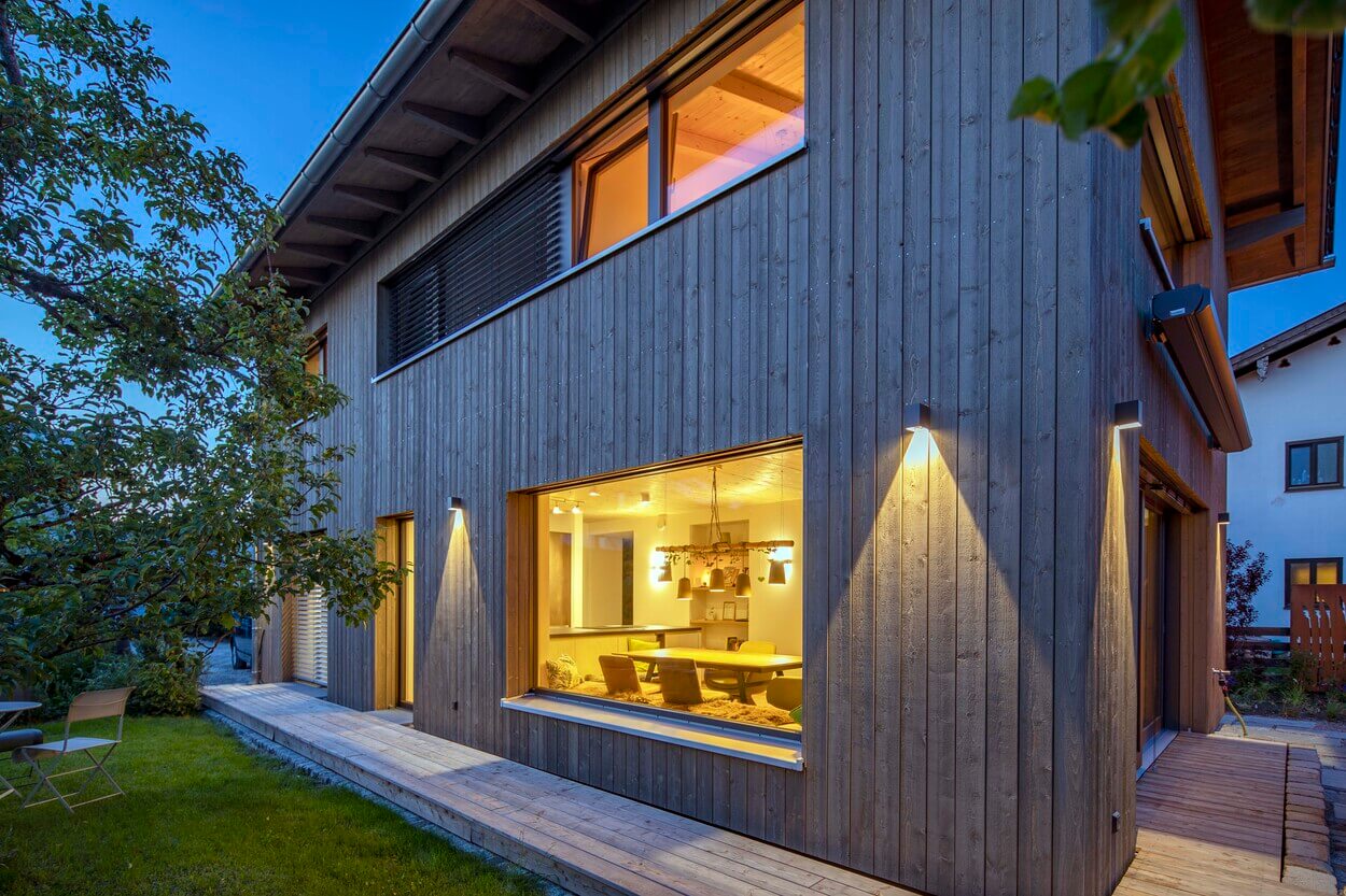 Ein modernes Holzhaus in Otterfing leuchtet bei Abenddämmerung mit erhellten Fenstern und einem gepflegten Garten.
