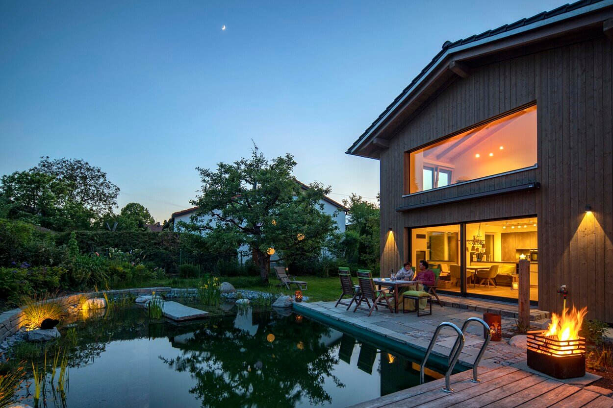 Ein modernes Holzhaus in Otterfing, beleuchtet bei Dämmerung, mit Menschen, die entspannt am Teich im Garten sitzen.