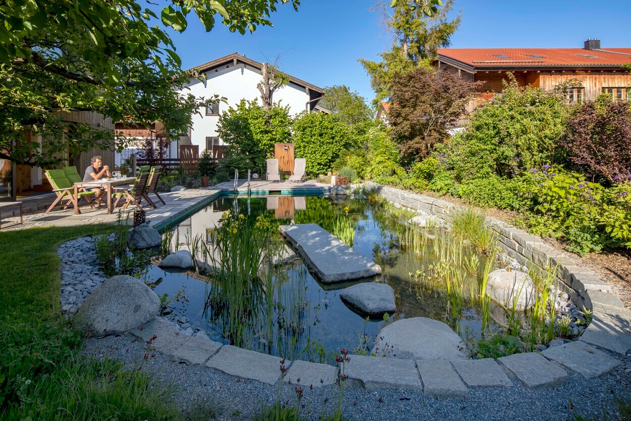 Ein Mann genießt seinen Morgenkaffee im sonnigen Garten in Otterfing, neben einem Teich und einem Holzhaus.