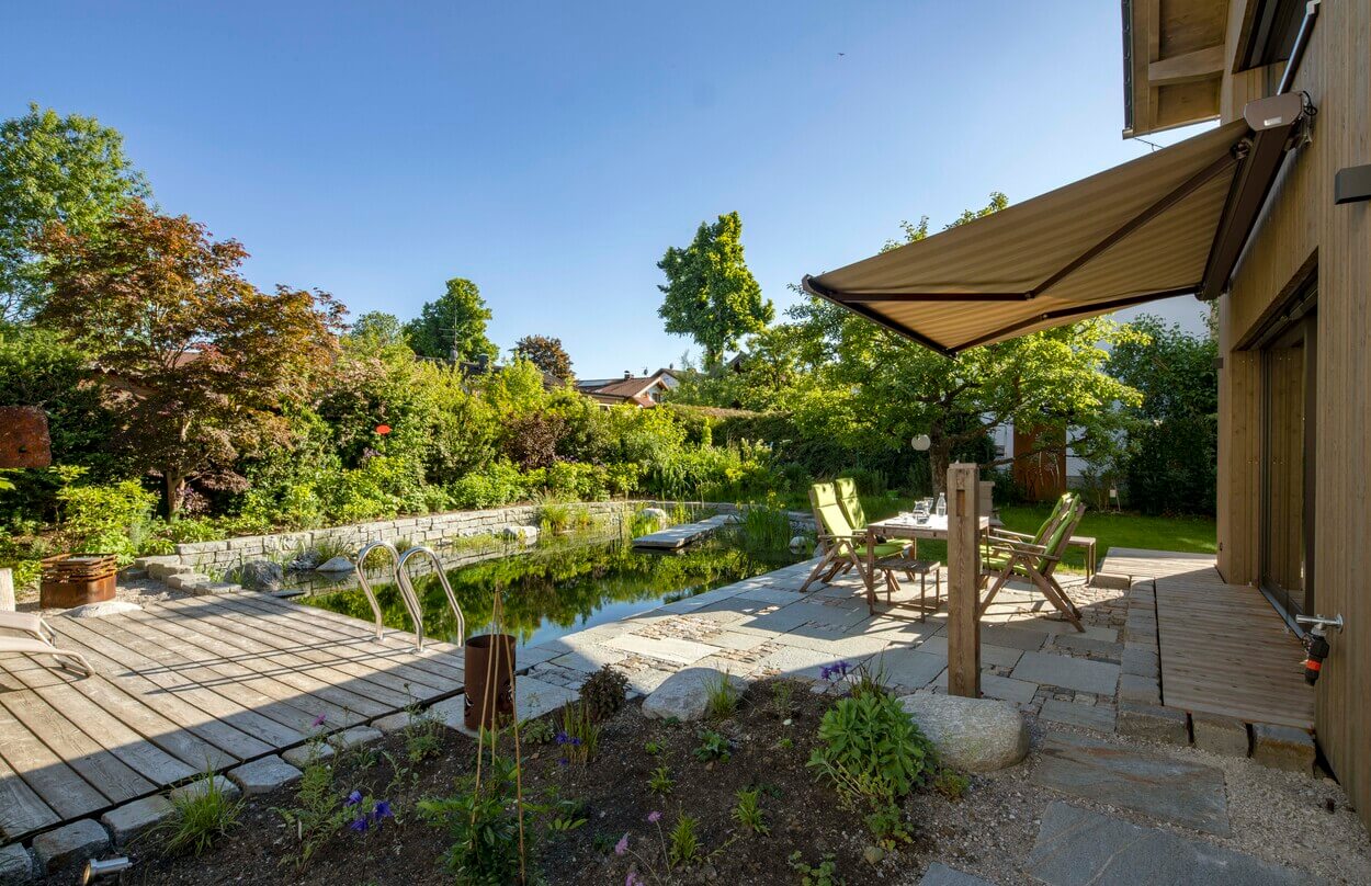 Ein Holzhaus in Otterfing mit Terrasse und Teich lädt unter blauem Himmel zum Entspannen im grünen Garten ein.