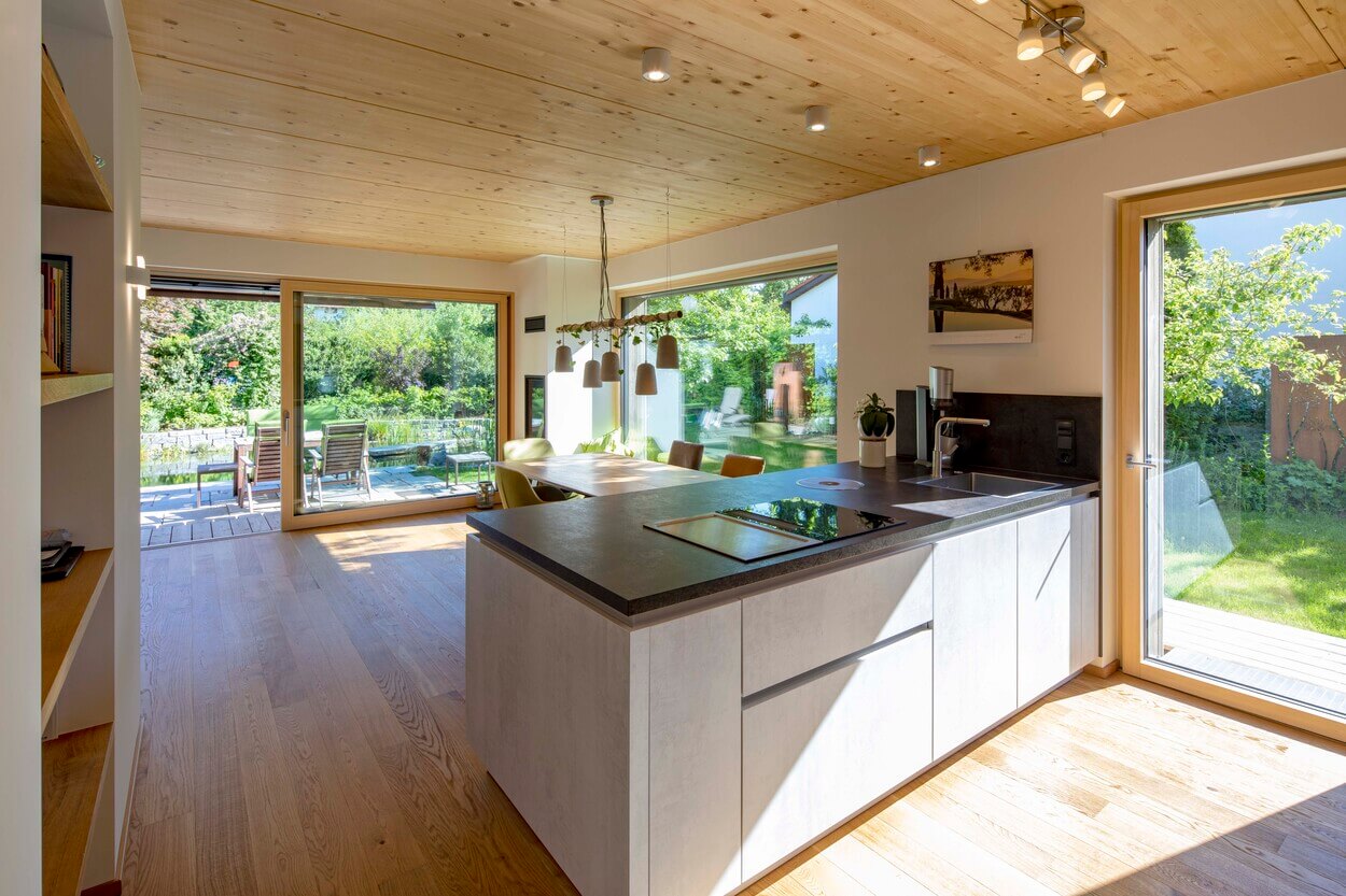 Eine moderne Küche im Holzhaus Otterfing zeigt durch große Fenstertüren einen sonnigen Garten mit Terrasse.