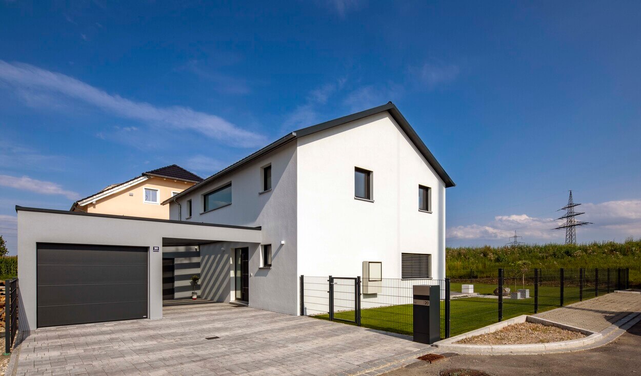 Moderne Außenansicht eines Passivhauses in Pliening mit gepflegtem Garten und strahlend blauem Himmel im Hintergrund.