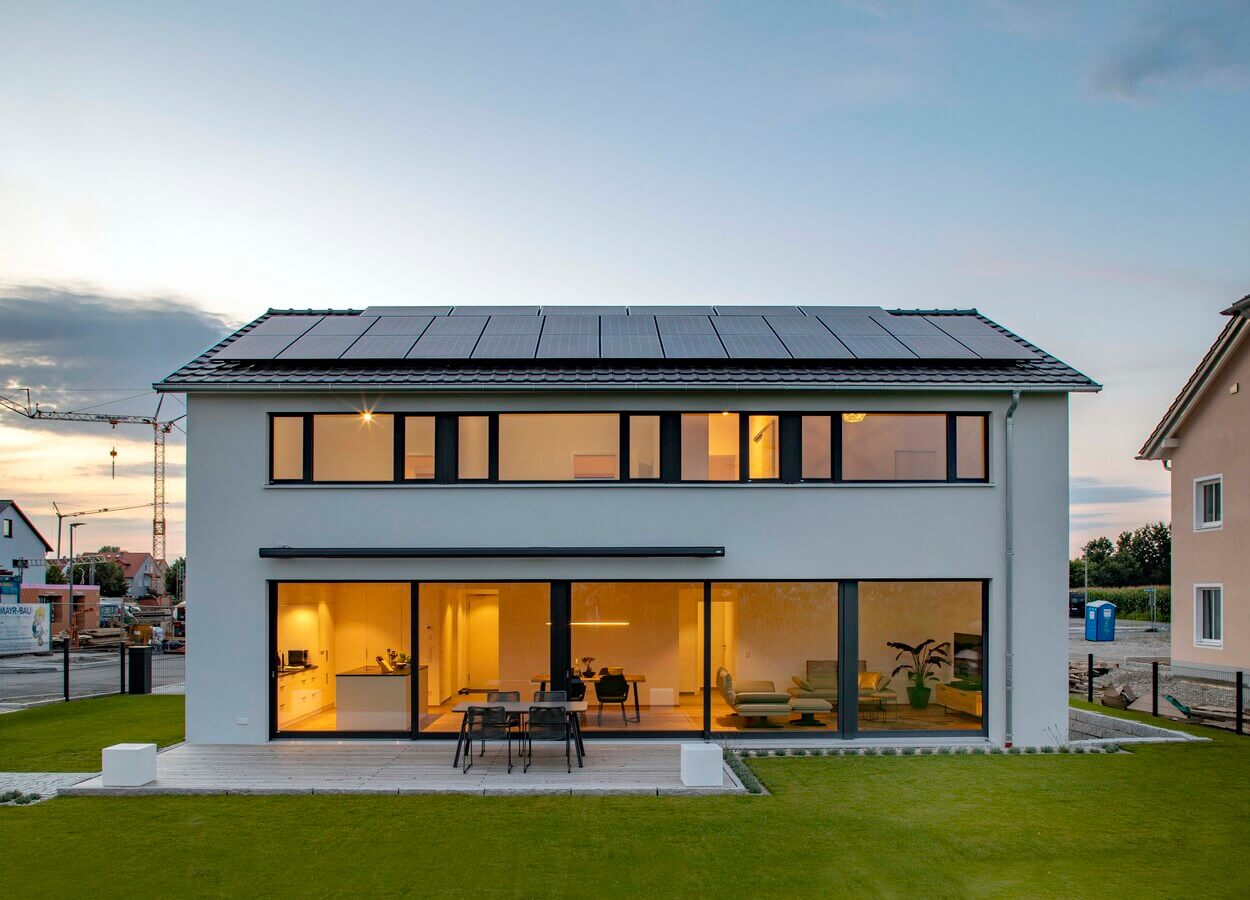 Ein modernes Passivhaus in Pliening mit Solarpaneelen, umgeben von gepflegtem Rasen, strahlt im Sonnenuntergang.