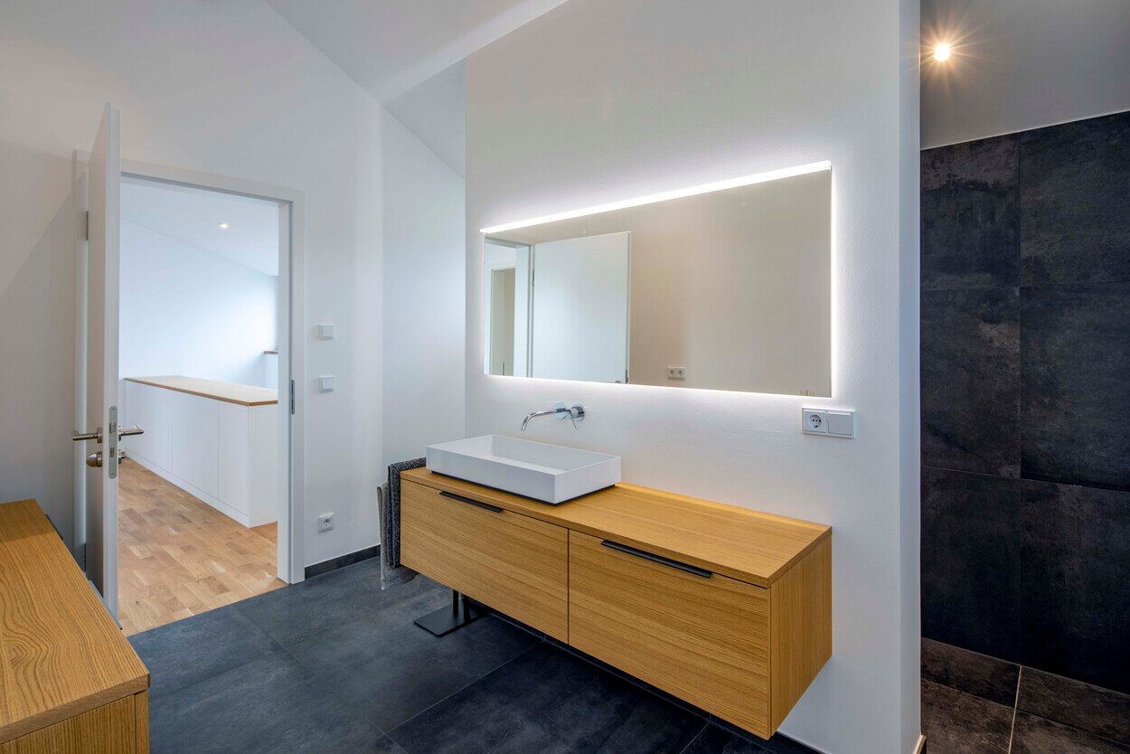 Ein modernes Badezimmer im Passivhaus Pliening mit hölzernem Waschtisch und beleuchtetem Spiegel sorgt für stilvolles Ambiente.
