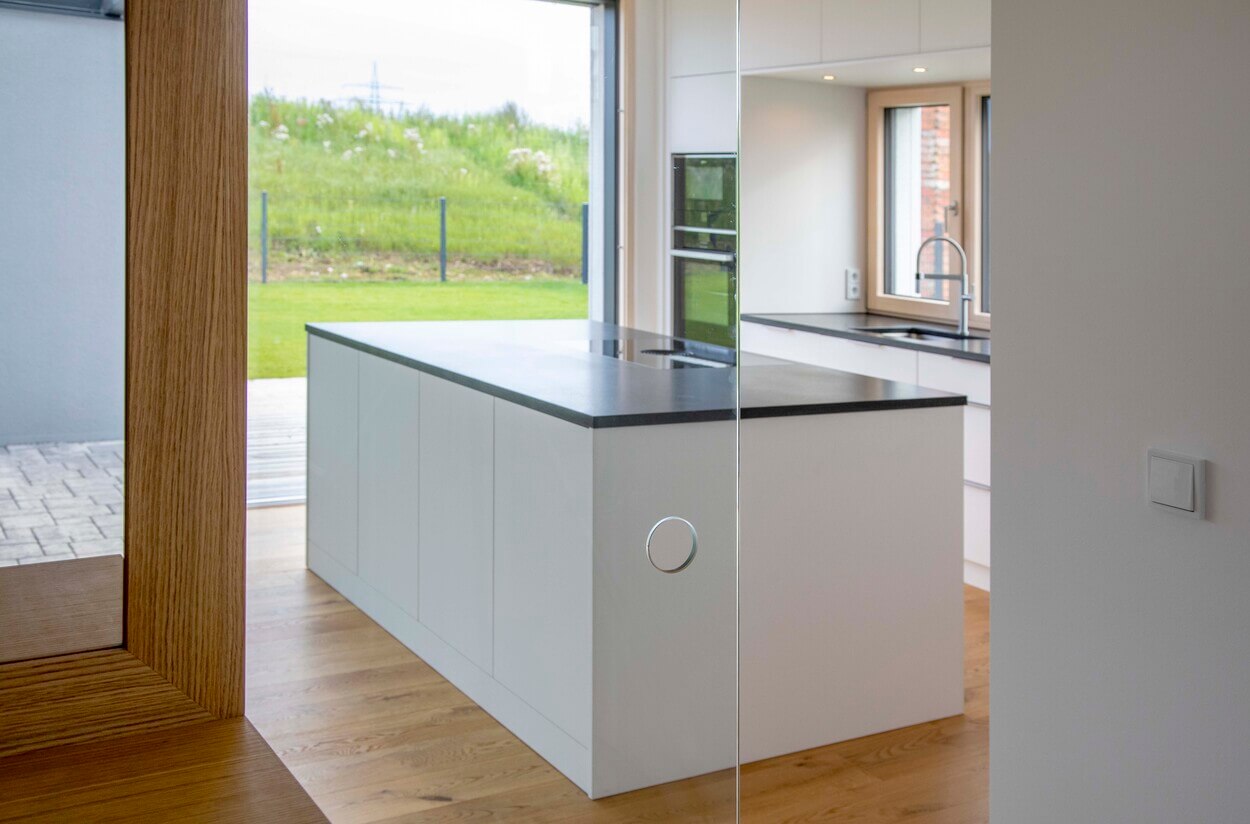 Die moderne Küche im Passivhaus Pliening bietet helle Schränke, eine schwarze Arbeitsplatte und einen herrlichen Blick ins Grüne.
