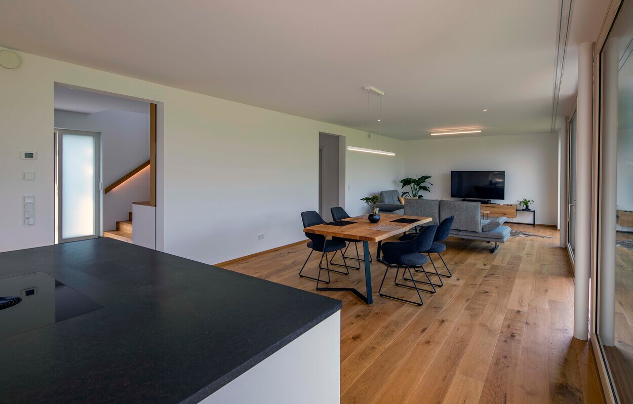 Der helle Wohnbereich im Passivhaus Pliening präsentiert einen modernen Esstisch und ein gemütliches Sofa vor einem großen Fernseher.