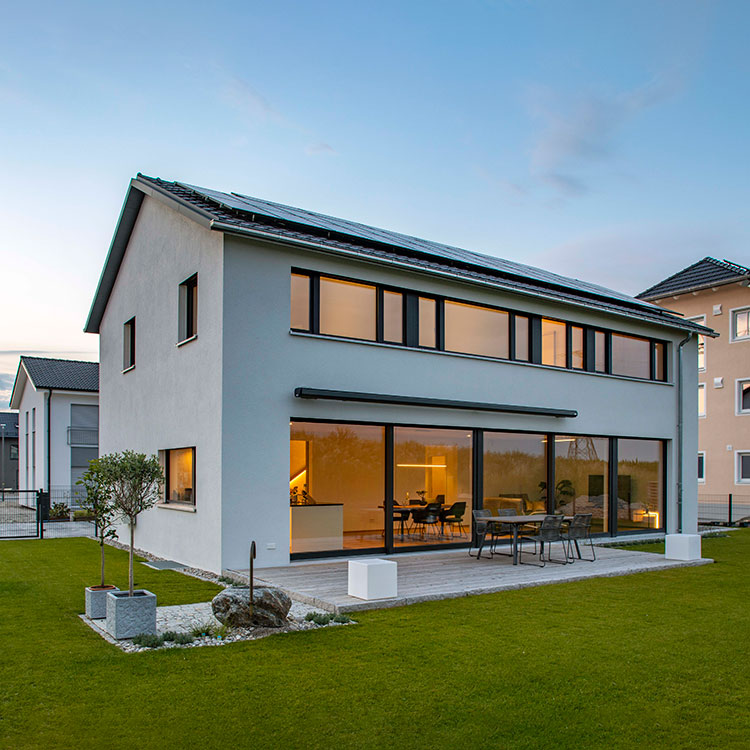 Ein modernes Passivhaus in Pliening mit großen Fenstern erstrahlt bei Abenddämmerung neben einem gepflegten Garten.