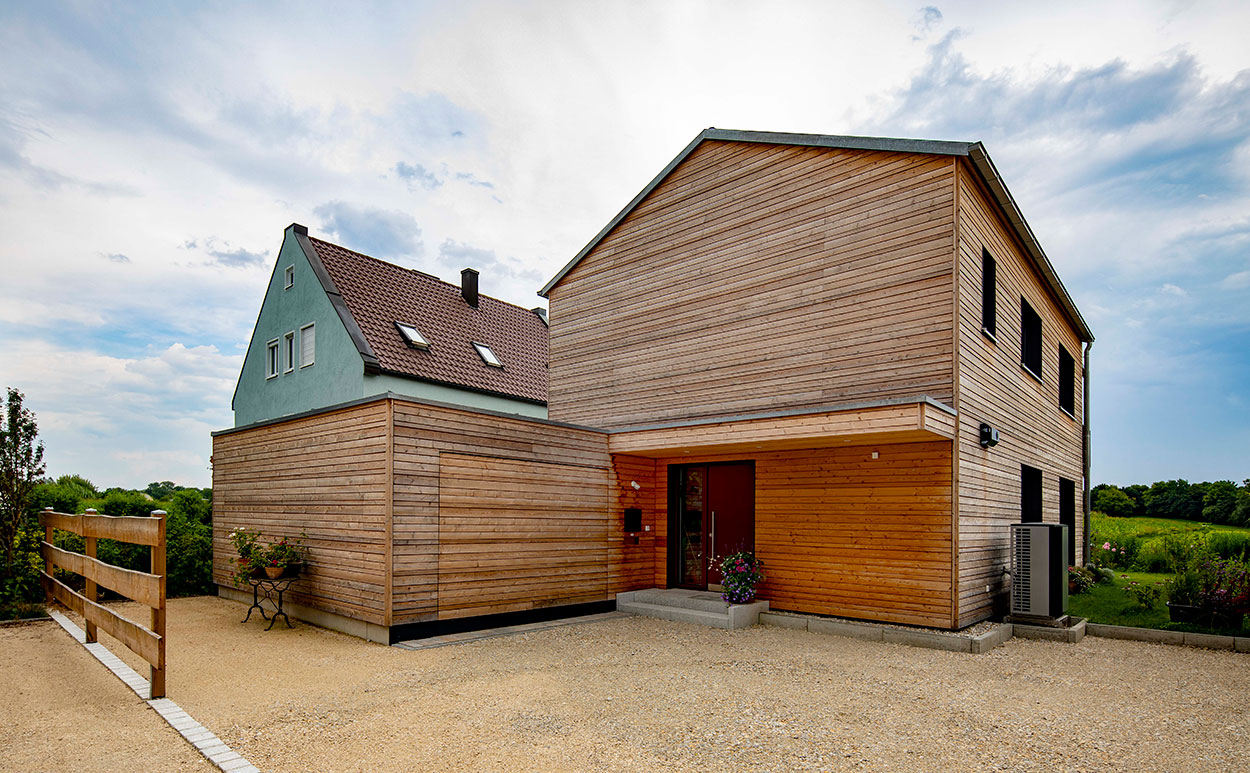Ein modernes Holzhaus in Dachau mit großzügigem Vorplatz und blühenden Pflanzen im Eingangsbereich unter bewölktem Himmel.