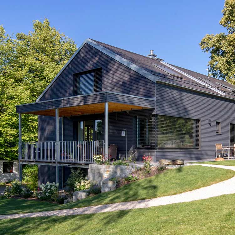 Ein modernes, dunkelgraues Haus mit großem Balkon steht idyllisch in einer grünen Landschaft in Glonn.