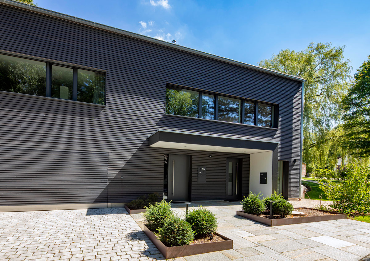 Der Eingang des modernen Holzhauses in Glonn ist von grünen Pflanzen und einem gepflasterten Weg stilvoll umrahmt.