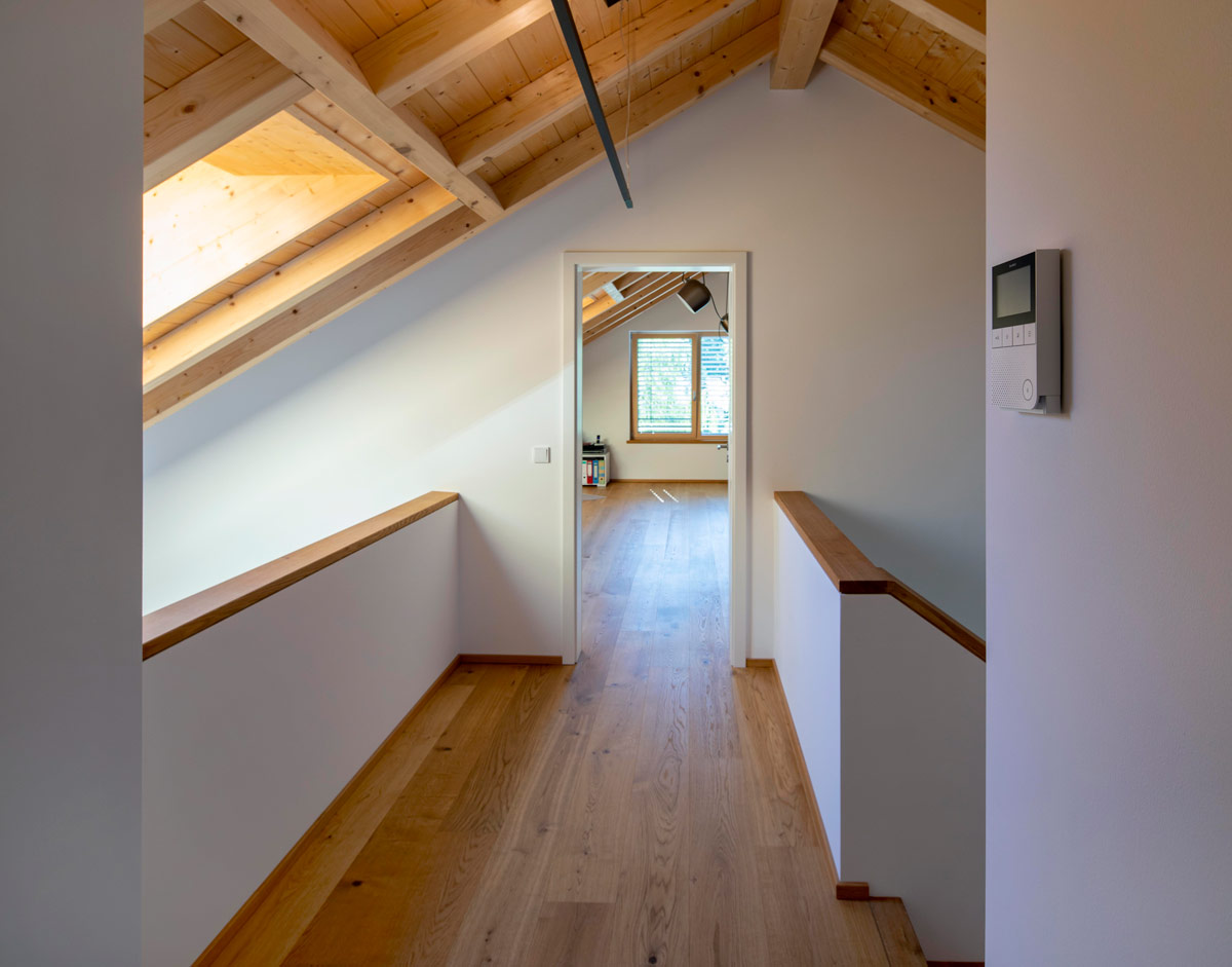Ein lichtdurchfluteter Flur im Dachgeschoss eines Holzhauses in Glonn führt zu einem gemütlichen Raum mit großen Dachfenstern.