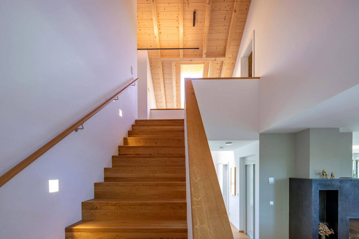 Eine hölzerne Treppe führt in einem modernen Holzhaus mit sichtbarem Dachstuhl nach oben, das Licht durchflutet.