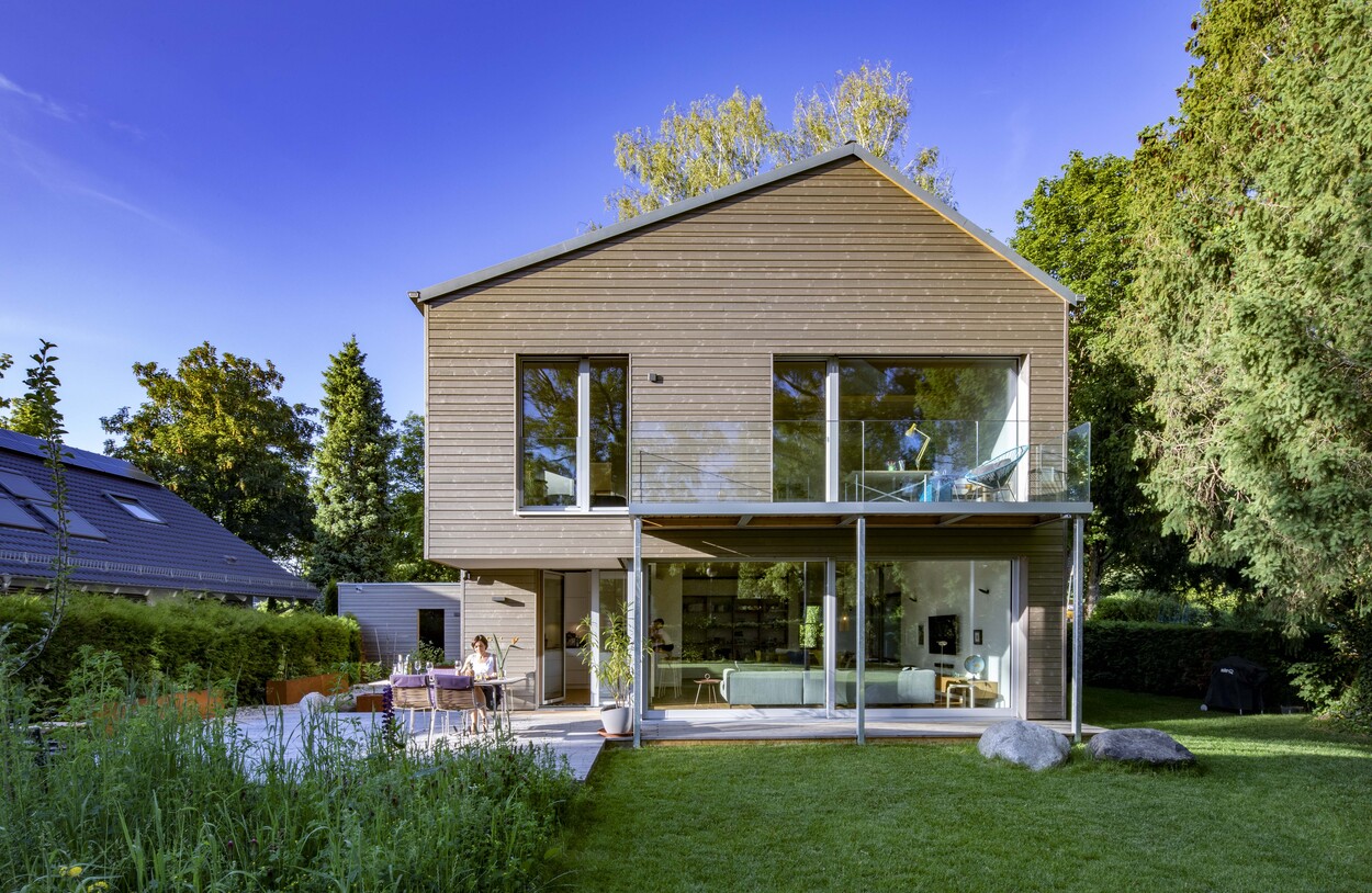 Ein modernes Holzhaus in Gröbenzell mit großen Fenstern steht in einem grünen Garten unter strahlend blauem Himmel.