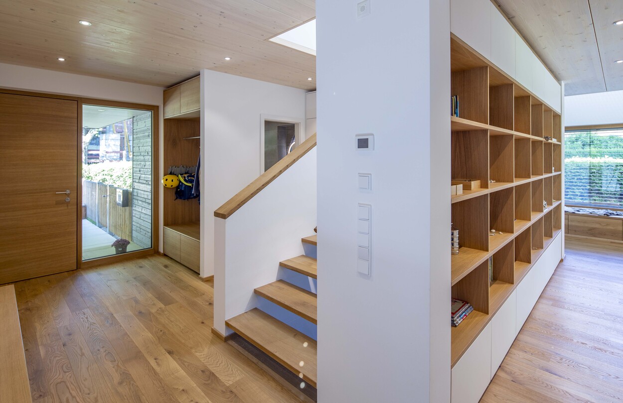 Ein modernes Holzhaus in Gröbenzell zeigt einen hellen Flur mit eleganten Holzregalen und einer stilvollen Treppe.